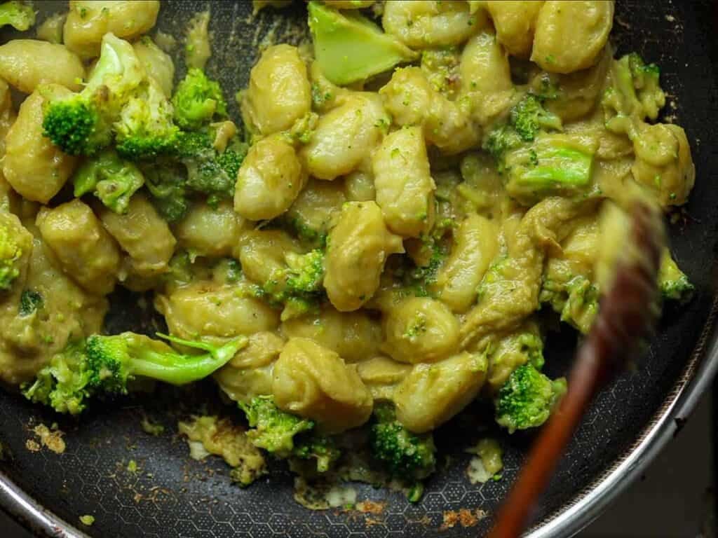 A frying pan filled with creamy gnocchi and broccoli florets, stirred with a wooden spoon. The gnocchi is coated in a thick, light brown sauce. The dish appears savory and freshly cooked.